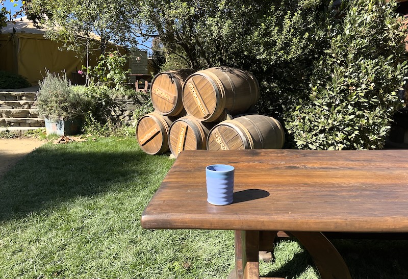 A blue pottery mug on a table. Barrels beyond.