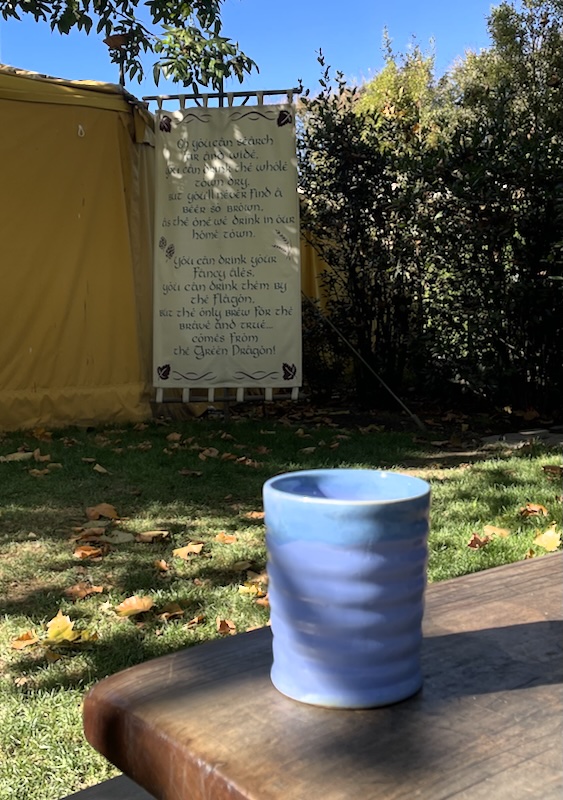 A blue pottery mug on a table. A banner beyond reads. Oh you can search far and wide. You can drink the whole town dry. But you'll never find a beer so brown as the one we drink in our home town. You can drink you fancy ales. You can drink them by the flagon. But the only brew for the brave and true... comes from the Green Dragon.
