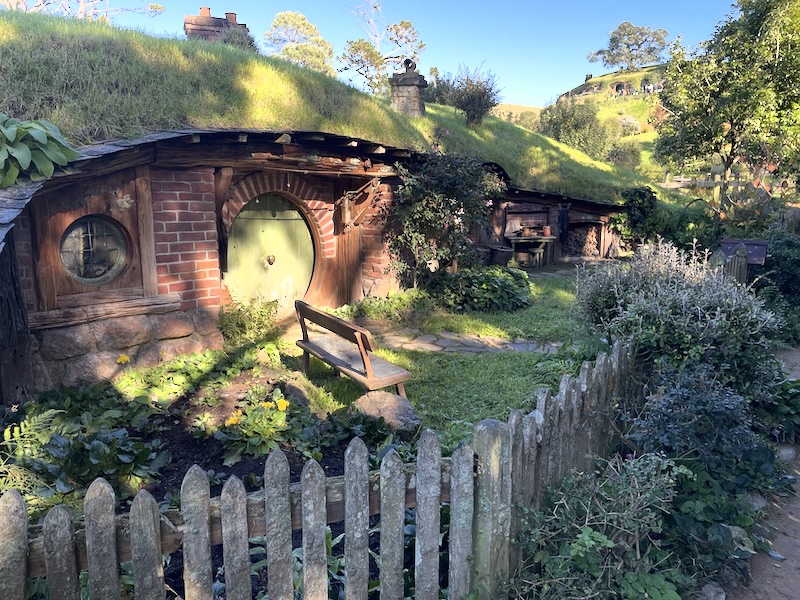 A hobbit hole with a yellow door. There is a bench in the front garden and a work bench to one side next to a log pile.