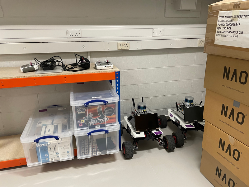 Two wheeled robots next to some racked shelving with Really Useful Boxes on the bottom layer. Carboard NAO boxes visible stacked to the right.