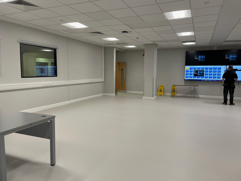 A large space with grey walls and a white floor. A desk is just visible on the right. A man stands at the far end in front of four screens.