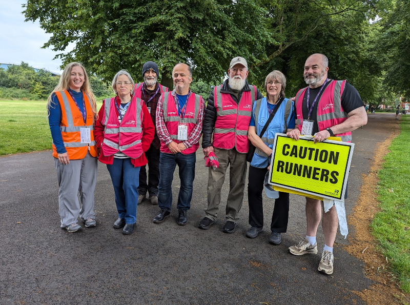 Picture of seven people in high vis jackets, most pink.  The rightmost person holds a Caution Runners sign.