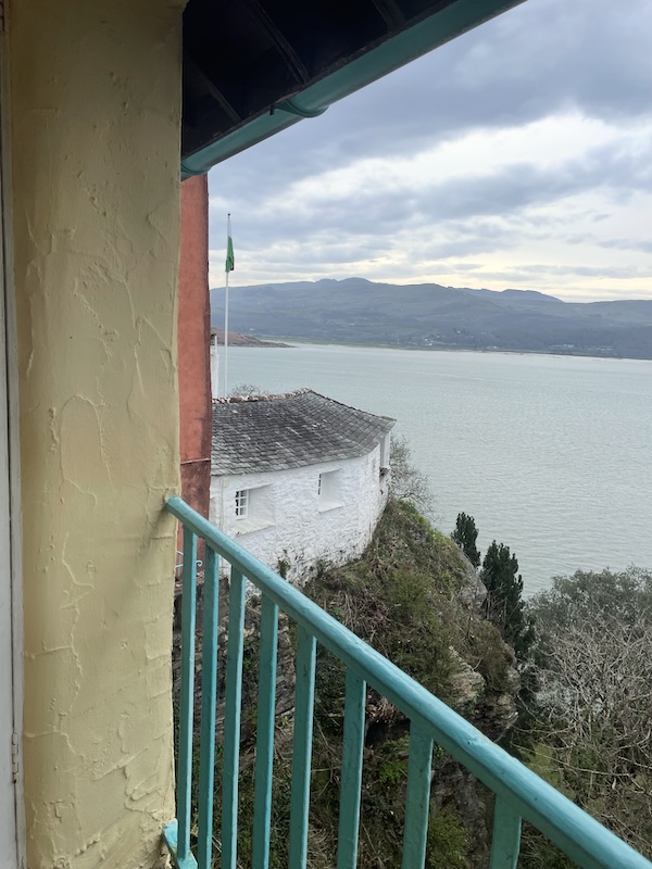 View from a window. Yellow plastered wall to the left. Blue railing. A cliff down to the sea with a white single storey building on top of it.