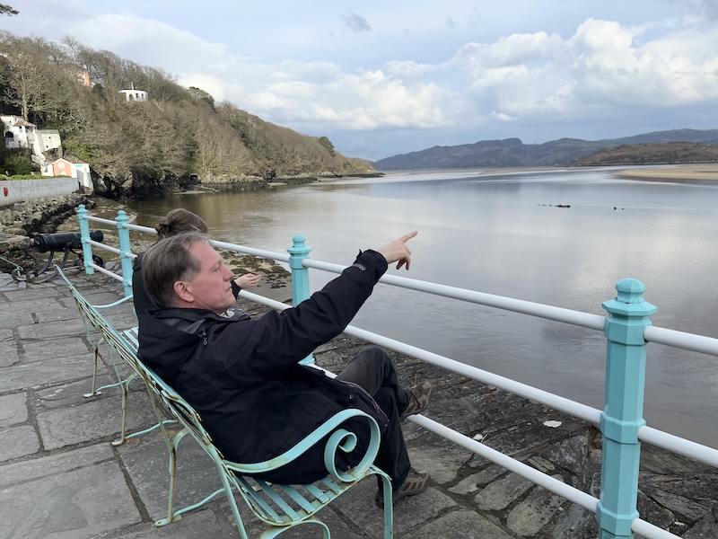 B. sitting on a bench before a railing with views over the estuary.
