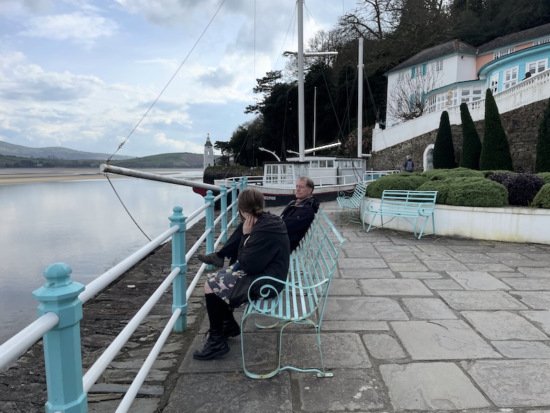 Marmalade Sparrow and B. sitting on a bench before a railing with views over the estuary. A stone boat is behind them.