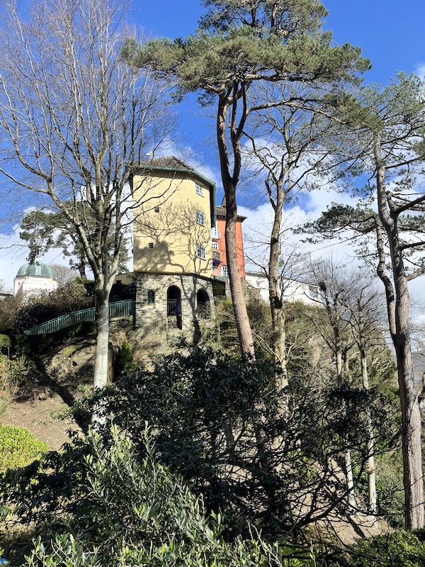 A view of a building on a cliff. The lowest level is bare stone arches. The top three levels are yellow plaster. Tall trees grow the height of the building.
