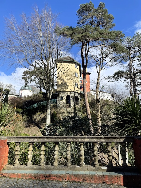 A view of a building on a cliff. The lowest level is bare stone arches. The top three levels are yellow plaster. Tall trees grow the height of the building. A stone balcony is at the front of the view.