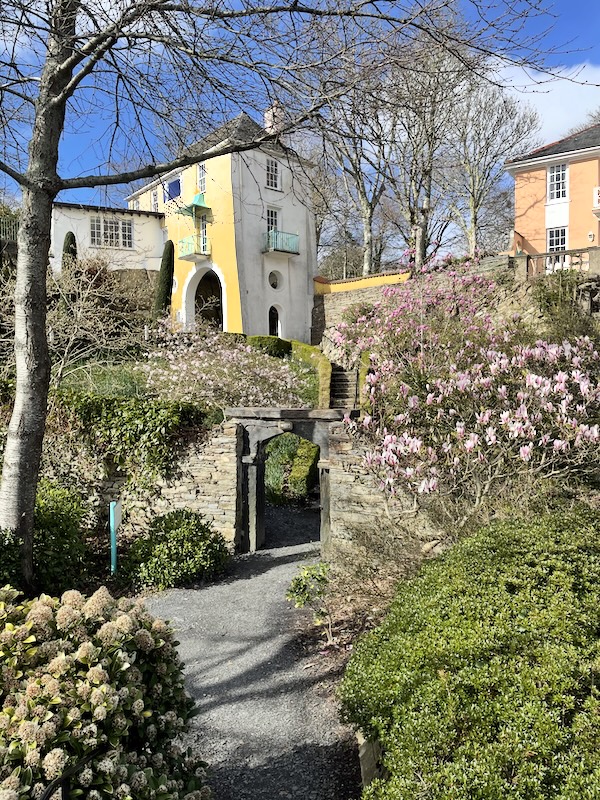 A view of a stone arch with a winding stair beyond leading to a yellow building with arches and balconies.
