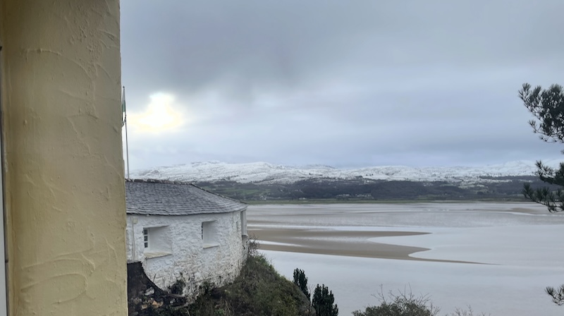 A view out across an estuary to snow covered mountains beyond. A white cottage is visible on the near cliff.