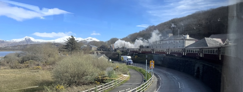 View of a steam train curving around on a raised track above a road. Blue skies and snow-capped mountains in the background.