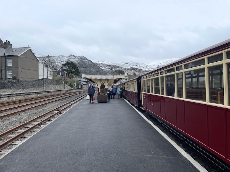 A station platform. Red carriages are pulled up on the left and snow-capped mountains dominate the background.