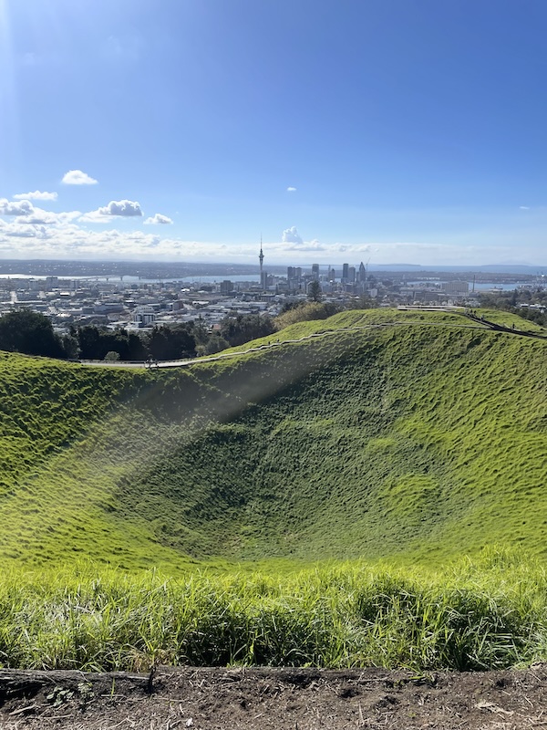 A pronounced grass covered dip in a hill top.  Beyond the Auckland skyline is visible, including a slender circular tower that tapers to a point.