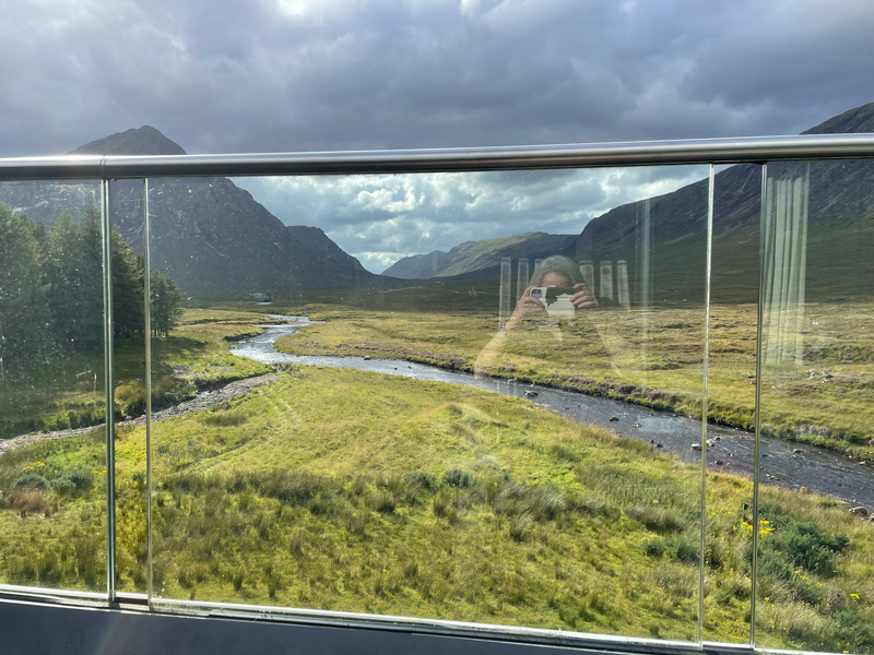 A view of mountains beyond a glass barrier. I can just be seen reflected in the glass, taking the photo.