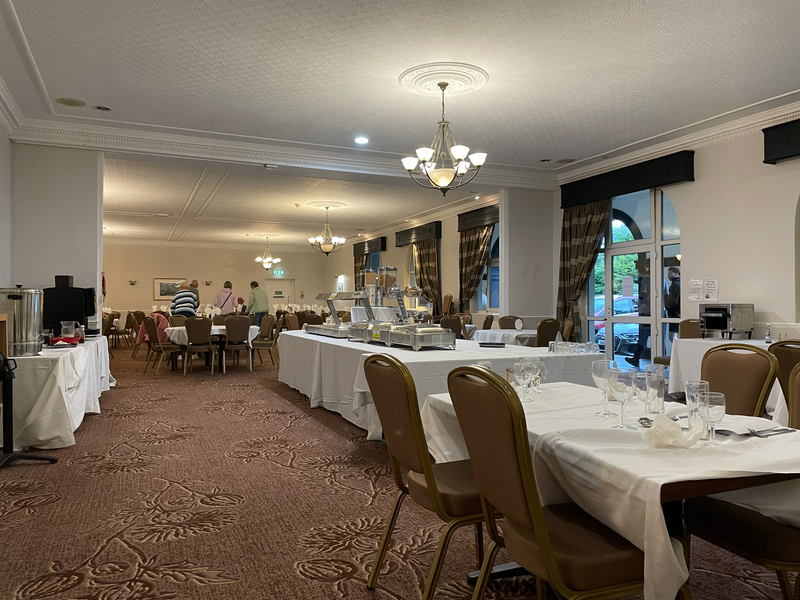 An empty rather beige room containing numerous empty tables and an empty breakfast buffet set up. A small number of people are seating themselves at one round table.