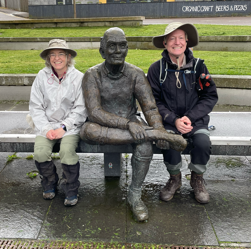 B. And I sitting either side of a statue of a man on a bench nursing one foot with its boot off.