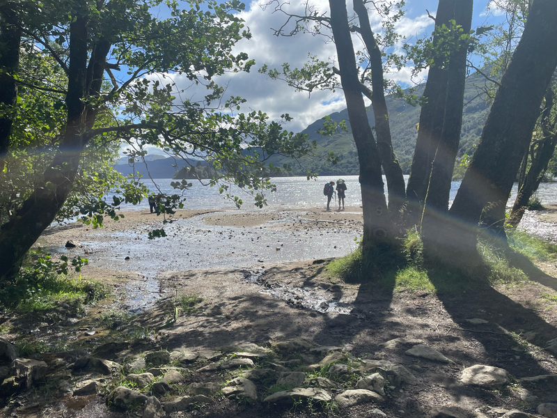 Two people standing on a pebbly beach by a loch in the sun with the mountains beyond.