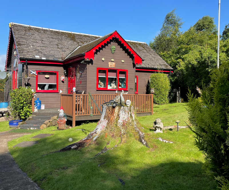 Wooden chalet with red decorative features. A man's head and arm emerges out of stump in the garden. Gargoyle like garden gnomes.