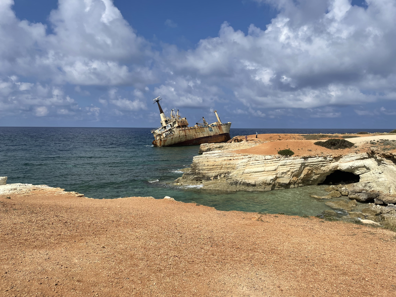 A mordern, metal cargo ship, lying at an angle next to a rocky coastline.  The sea is bluey green. The ship is streaked with rus a similar colour to the sand on top of the rocks.