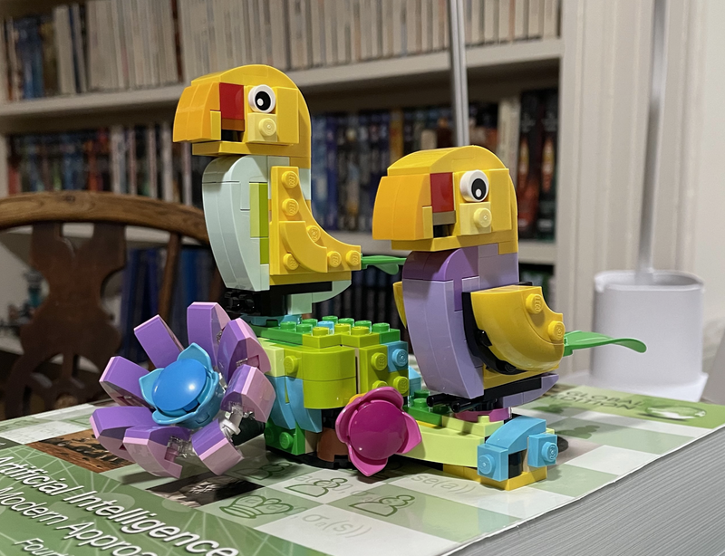 Two yellow lego birds, one with a blue breast and one with a purple breast sitting on a flower.