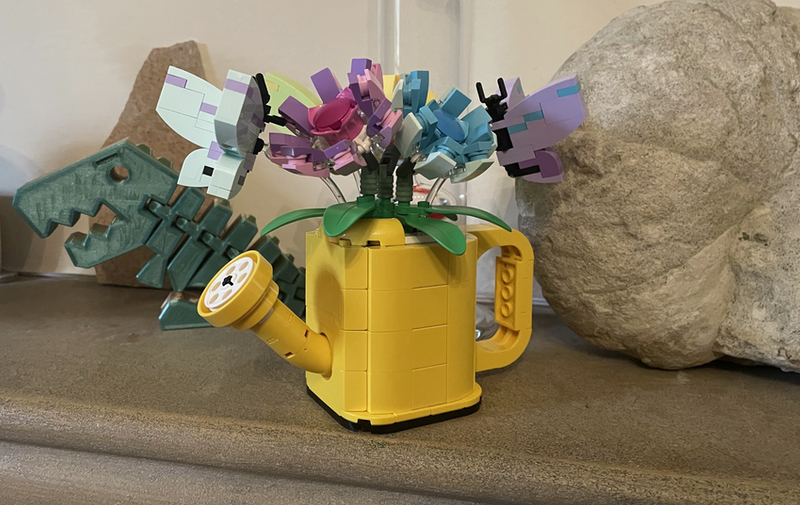 A yellow lego watering can with flowers in it and butterflies on the flowers.
