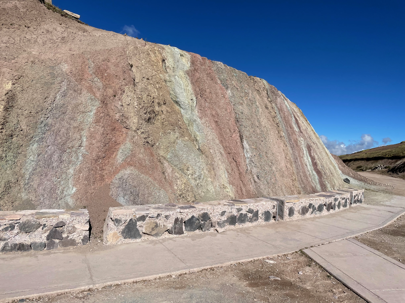 A paved walkway, edged by rock and then a cut away bit of hillside revealin red, green and white stripes