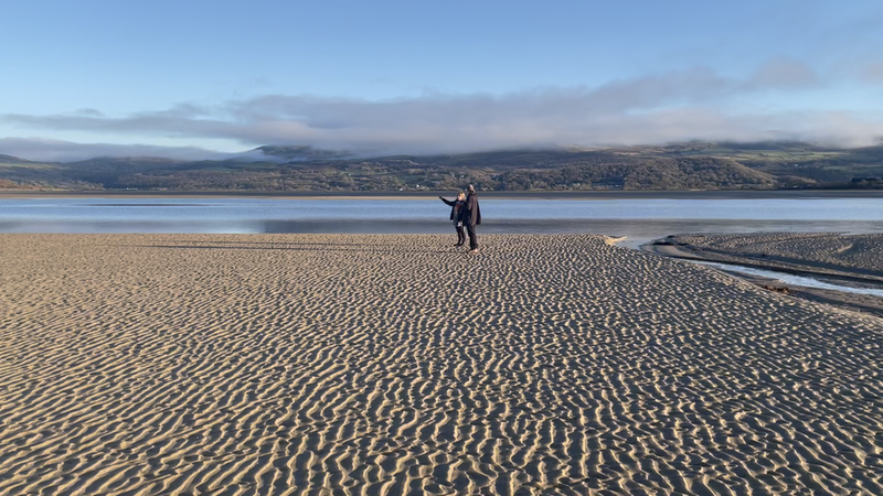 Two people on a sandy beach.  Hills visible across the estuary.
