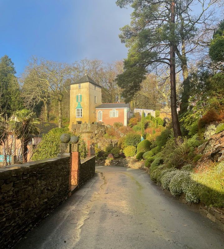 A yellow tower with a red building next to it.  A road leads to it with a steep bank and a tree on one side and a wall with a gate on the other.  The sky is a threatening blue, but sunlight is on the building.