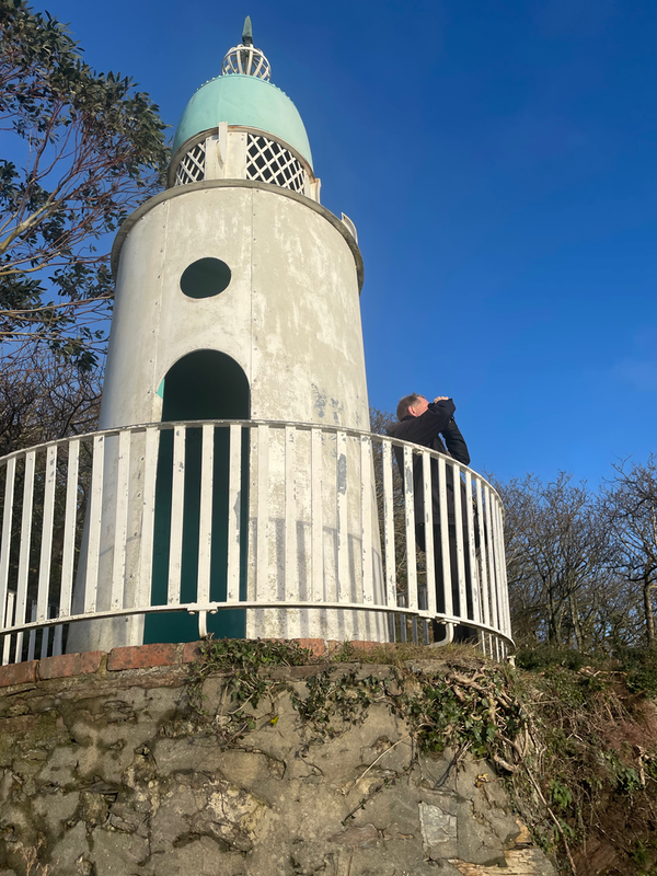 A man standing on a balcony around a white lighthouse.  He is looking through binoculars.