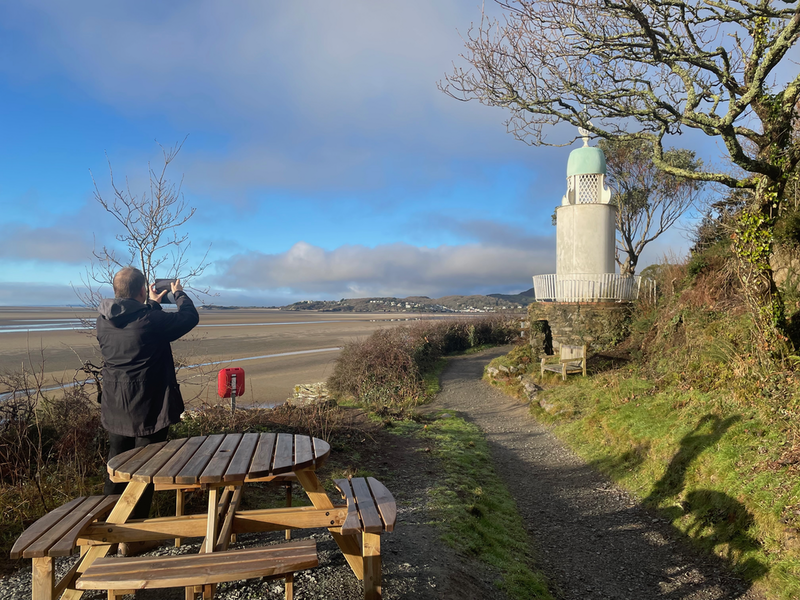 A white lighthouse with a blue top against beach and blue sky.  A man stands by a picnic table to the left, facing away from us, taking a photo.
