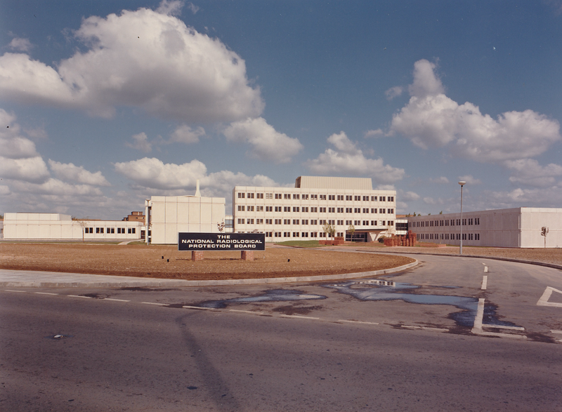Colour photo of a white rectangular building in bare grounds. A sign announces The National Radiological Protection Board.