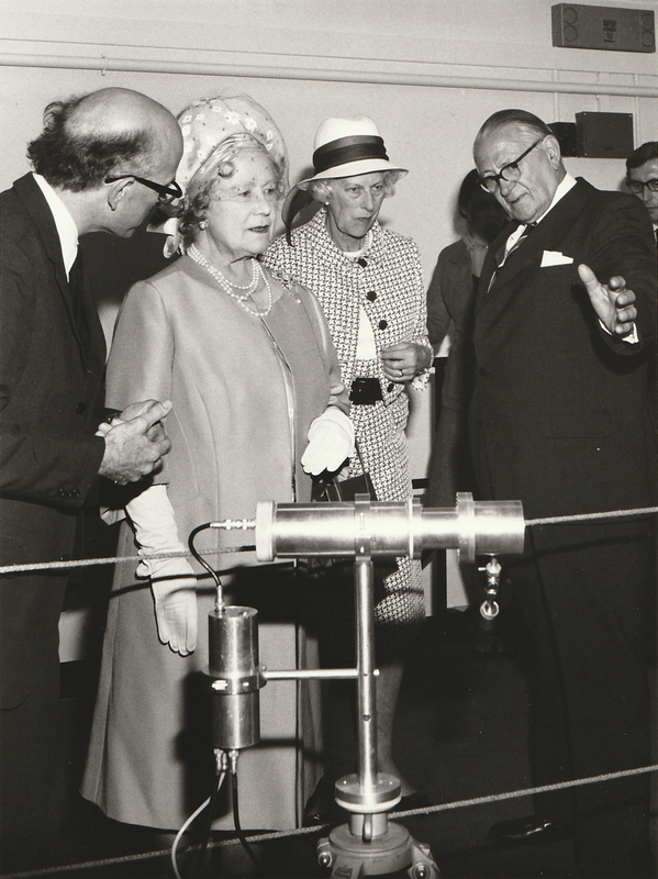 The Queen Mum in front of some kind of cyclindrical device. A lady in a tweedy looking outfit stands behind her. They are flanked by two men in suits. The nearest, with his back to us, is bald.