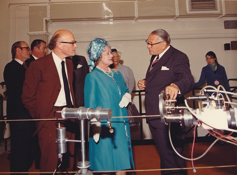 Colour photo of the Queen Mum in a blud coat and hat. In front of the presumed neutron generator, flanked by two men in suits. The bald man on the right's suit is brown.