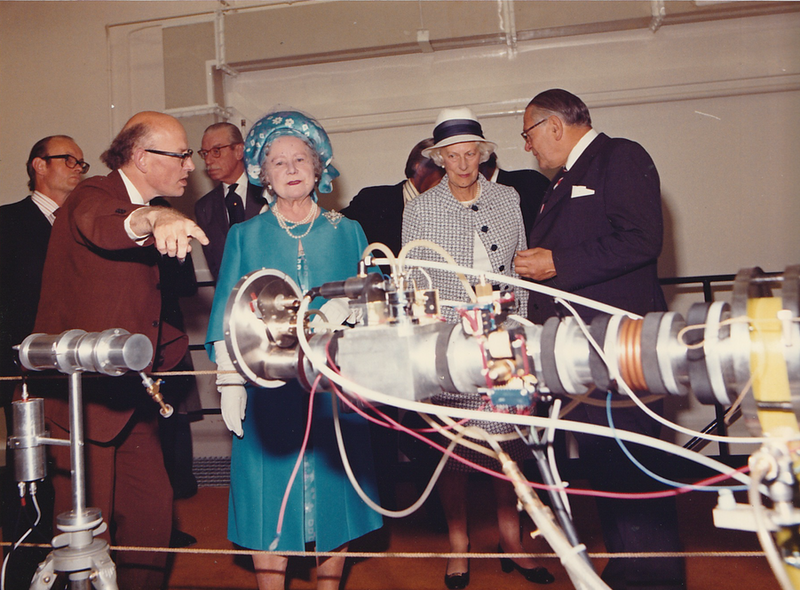 Color photo of the Queen Mum. The bald man in the brown suit is pointing at the presumed neutron generator.