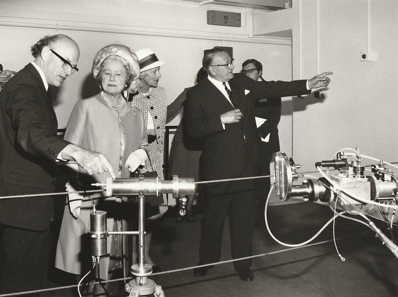 Black and white photo of the Queen Mum. The bald man in the brown suit is pointing at the presumed neutron generator. Behind them the other suited man points out of shot.