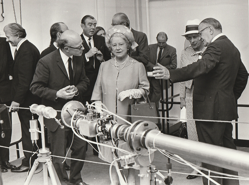 Black and White photo. They are still looking at the neutron genrator. Bald man seems like he's paying careful attention.