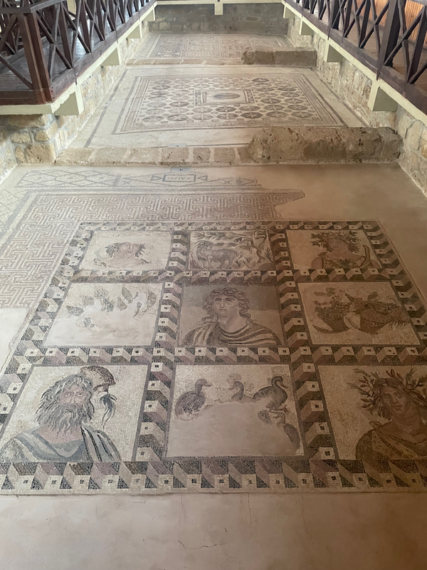 View along the remains of three small square rooms with mosaic floors in each. The closest is most elaborate with a square of nine images, those in the corners and center of the heads of people.