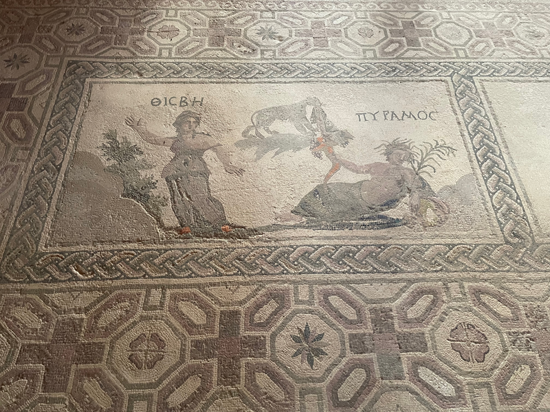 A mosaic with a geometric border and a woman who appears to be fleeing. A man reclines holding some kind of horn. What could be a lion sort of floats between them with a garment in its mouth. Words tell us the woman is Thisbee (in greek lettering) and the man Pyramos (in greek lettering)