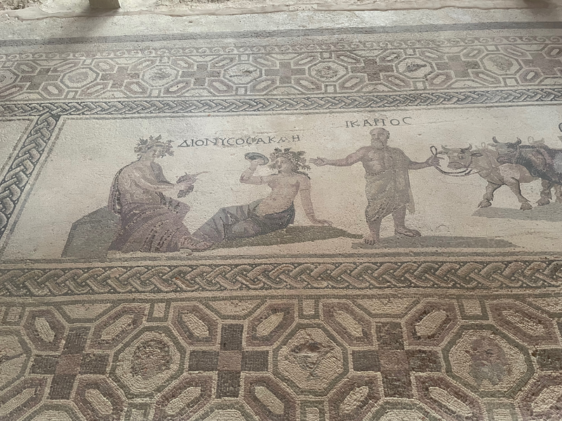 Half fof a mosaic showing a garlanded man with possibly a drinking horn and a woman with a plate.  Another man is leading oxen.  Over the first man and woman is the work Dionysos Akah (in greek) and above the other man is Ikarios