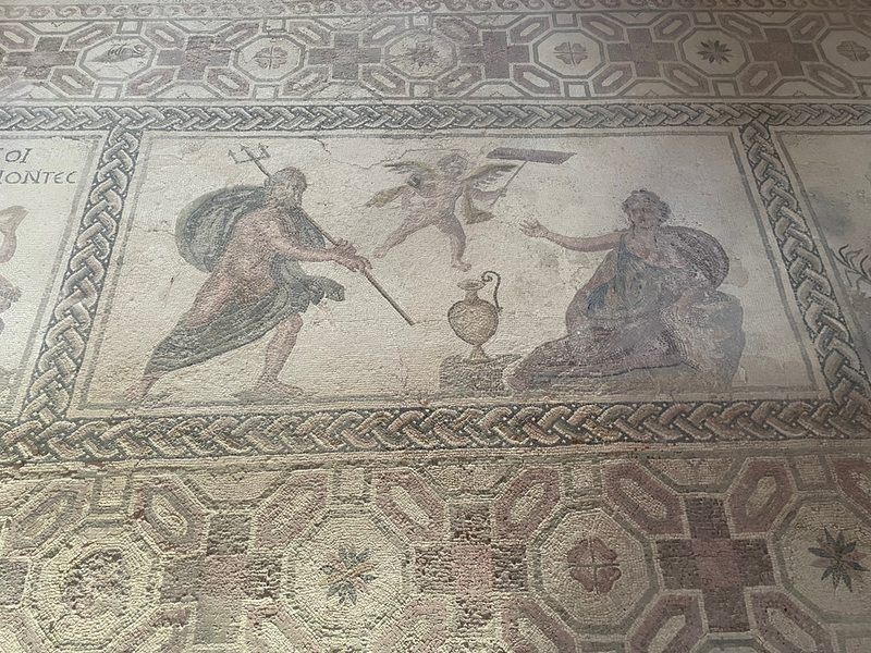 A mosaic of a man with a trident, a seated lady holding out her hand and a small winged person holding a stick with some kind of plank on the end.