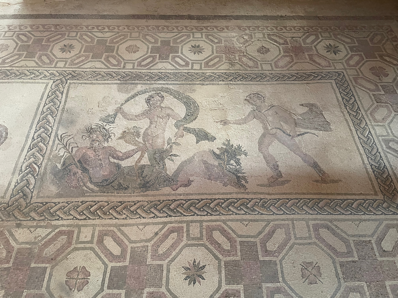 A mosaic of a reclining garlanded man with a horn, a naked lady with a long floating scarf, and a naked man with small bow.