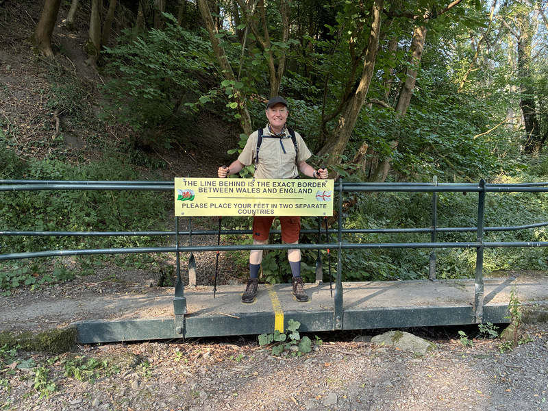 A man stands proudly behind a sign that reads The Line Behind is the Exact Border between Wales and England. Please Place your feet in two Separate Countries.