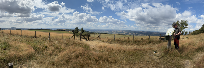 Panorama shot showing mostly dry grass and fencing, but there are patchwork fields beyond and a man to one side trying to consult his phone in the shade of an info-board.