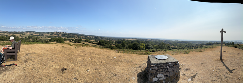 Panorama: Man sitting on bench, landmark info stone, signpost - all with patchwork of fields beyond.