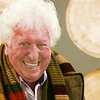 An older Tom Baker with white hair wearing the fourth Doctor's scarf with Tardis roundels in the background.