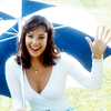 Nicola Bryant in a white top and a blue and white umbrella waving at the camera.