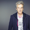 Publicity shot of the Twelfth Doctor waist up against a grey background.  Looking to camera in T-shirt and hoodies.