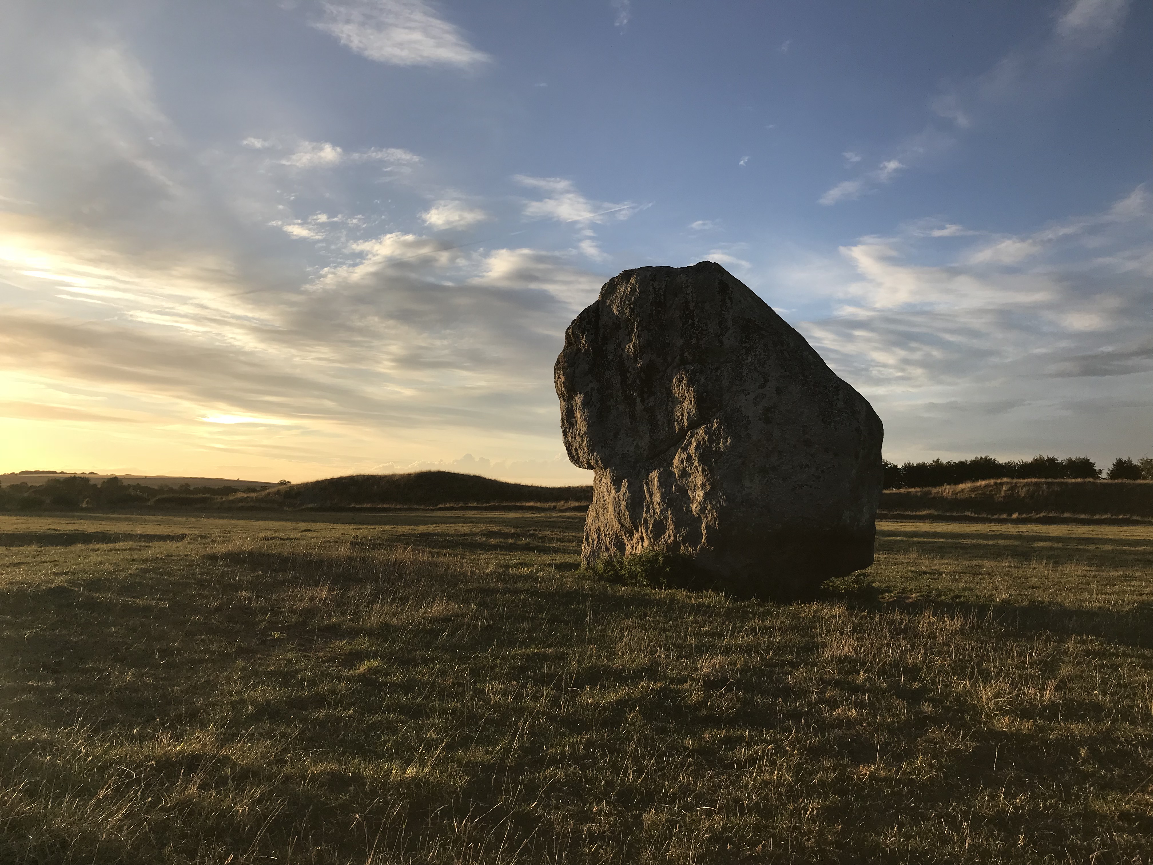 A large lump-like standing stone, alone in the grass, a bank is visible beyond if you know what to look for. It is sunset.