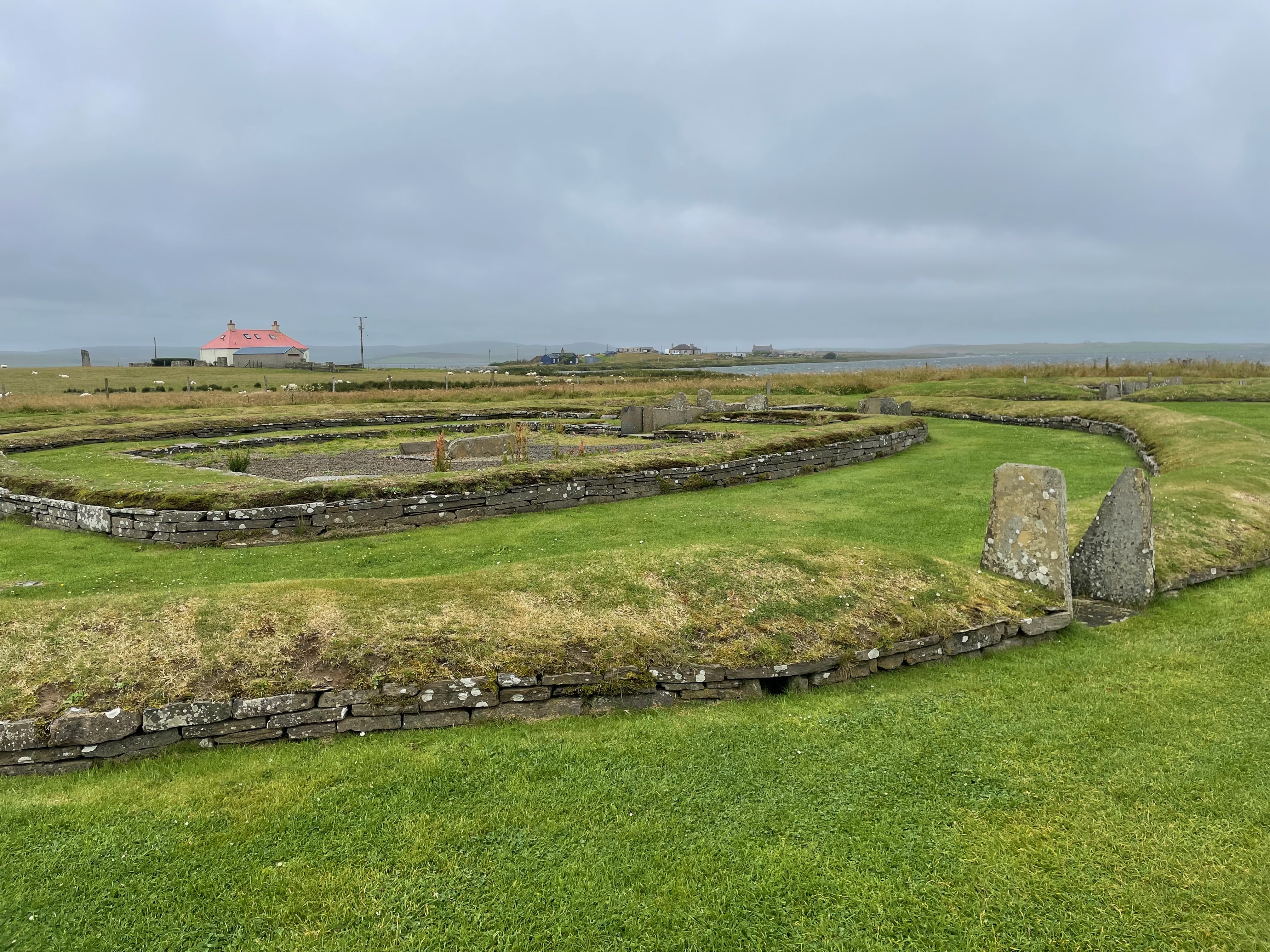 The remains of walls made from flat stones in a rough circle with grass on top, surrounded further similar remains.  An entrance with two upright flat stones is to the right.  Beyond is a view of a white building with a red, roof, water, and hills in the mist.