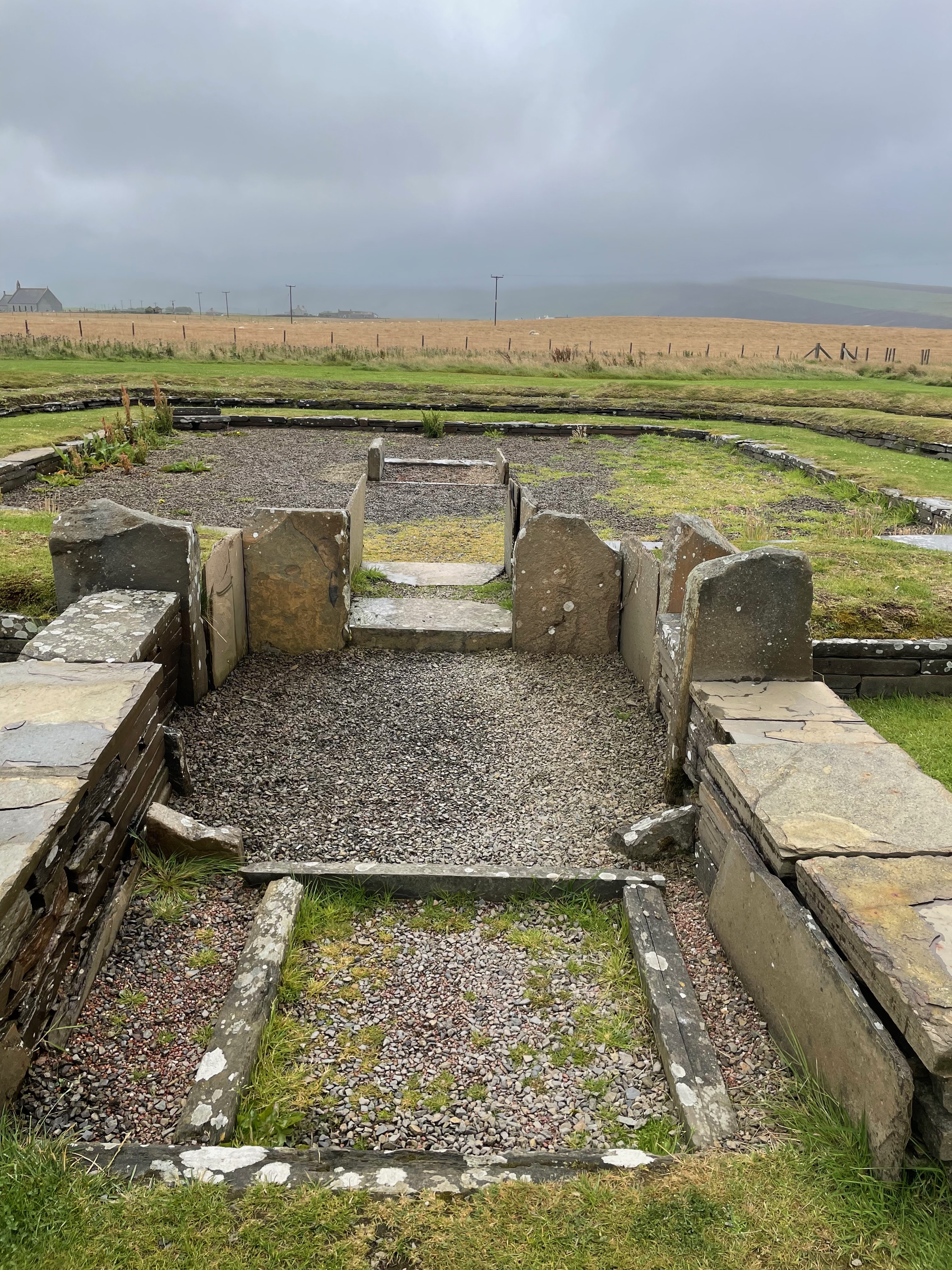 The entrance way to what looks like a super-low arena with only two tiers. The entrance is surrounded by thin upright stones, about knee height but still higher than the structure beyond. There is some kind of hearth in the centre of the arena structure.