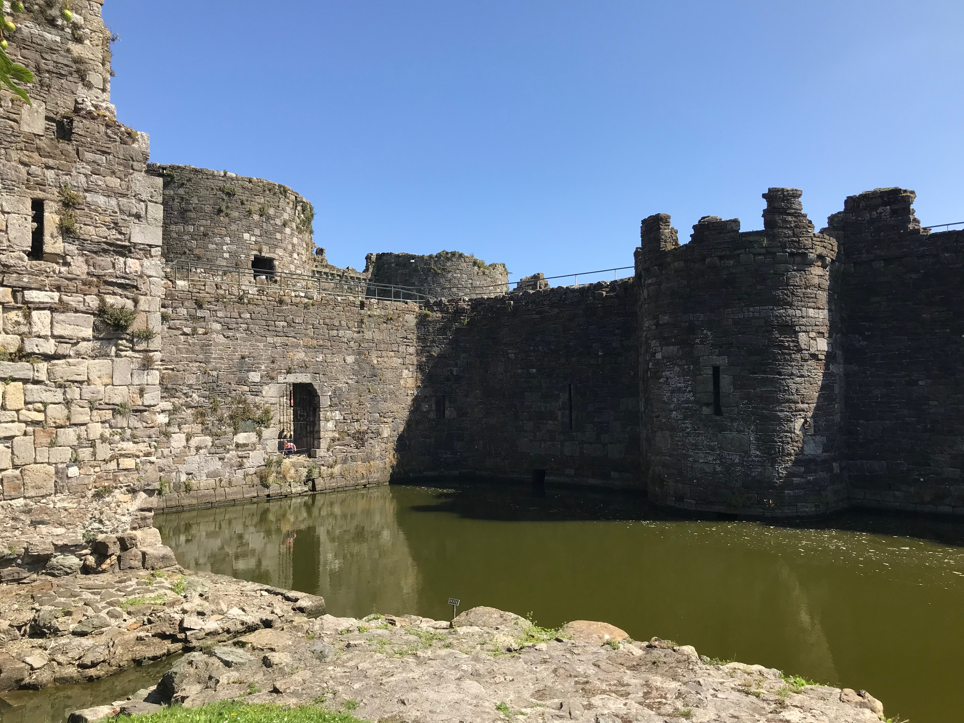 Two stone walls, one with a turret in the centre surrounding a pool of water.  To the front a third side is a stone walkway.